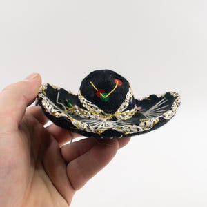 May include: A small black sombrero with a rounded crown and a broad, flat brim. The brim is trimmed with gold and silver, and features colourful embroidered details. A traditional Mexican hat design.