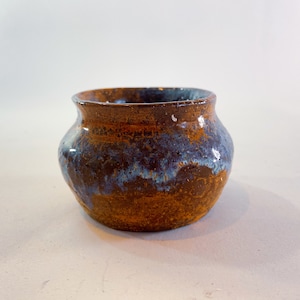 May include: A small, round, brown ceramic pot with a blue and white glaze. The pot has a slightly flared rim and a rounded bottom.