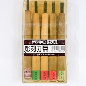 May include: A set of five wood-handled carving tools in a clear plastic case. The tools have dark green blades and are labeled with Japanese text. The packaging includes the text "Sakura Cray-Pas Hanga" and "Chokukuto 5B".