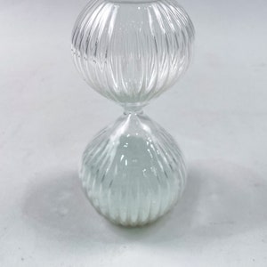 May include: A clear glass hourglass with a ribbed design. The hourglass is filled with white sand.