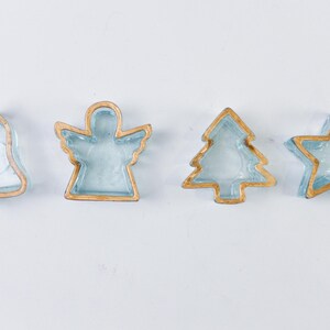 May include: A set of four light blue glass ornaments with gold-coloured edges, shaped like a bell, an angel, a Christmas tree, and a star. Each piece features a translucent blue centre surrounded by a metallic gold-toned border outline.