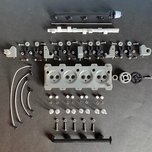 May include: A disassembled model engine with various parts, including a cylinder head, pistons, connecting rods, and a crankshaft. The engine is made of metal and plastic and is painted in a silver and black colour scheme.