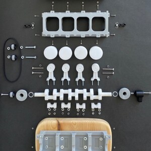 May include: A disassembled model of a four-cylinder engine with various parts laid out on a black surface. The parts are made of white and grey plastic and include pistons, connecting rods, a crankshaft, and a timing belt. The engine is mounted on a wooden base.