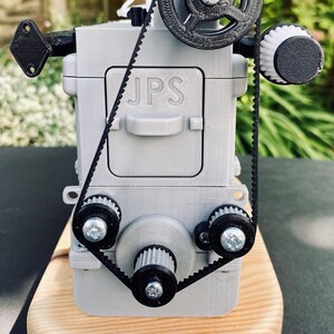 May include: A 3D printed model of a small engine with a black belt and gears. The engine is grey and has the letters "JPS" printed on the side. The engine is sitting on a wooden base.