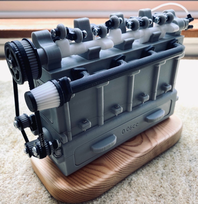 May include: A 3D printed model of a small engine with a black belt and a white air filter. The engine is mounted on a wooden base and has a label that reads "0.06cc".