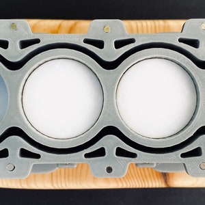 May include: A grey 3D printed model of a four-cylinder engine with four white cylinders. The engine is mounted on a wooden base.