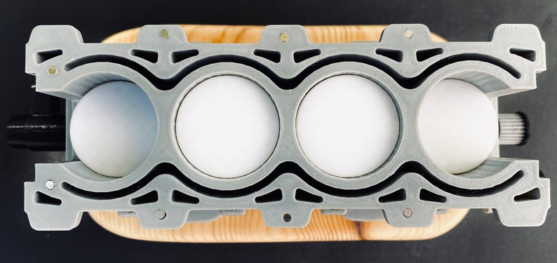 May include: A grey 3D printed model of a four-cylinder engine with four white cylinders. The engine is mounted on a wooden base.