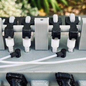 May include: A close-up of a grey 3D printed model of a V8 engine with black pistons, white spark plugs, and a black belt.