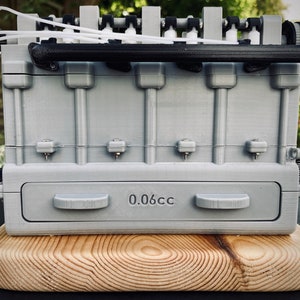 May include: A 3D printed model of a small engine with a grey finish. The engine has a black handle and a label that reads "0.06cc". The engine is sitting on a wooden base.