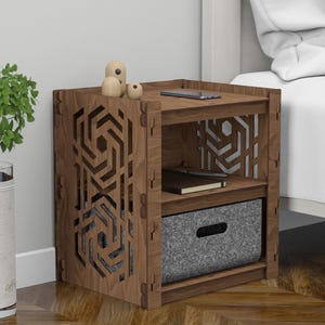Può includere: Un comodino in legno con un design geometrico sul davanti. Il comodino ha due ripiani e un cassetto. Il cassetto è grigio e ha una maniglia. Il comodino è su un pavimento in legno.