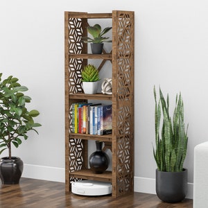 May include: A brown wooden bookshelf with five shelves and a geometric pattern on the sides. The shelves are filled with books, plants, and a vase. A white robot vacuum cleaner is on the bottom shelf.