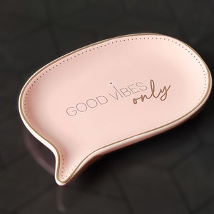May include: A pink, speech bubble-shaped trinket dish with gold trim and the words "GOOD VIBES only" in gold lettering. The dish has a dotted border and sits on a dark, geometric patterned surface.