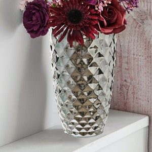 May include: A decorative silver vase with a diamond pattern, filled with artificial flowers. The bouquet features burgundy gerberas, roses, and other flowers in shades of purple and pink. The vase is on a white shelf.