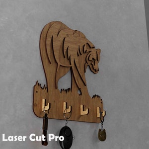 May include: Wooden bear-shaped key holder with four hooks. The bear is cut from wood and has a detailed design. The key holder is mounted on a gray wall, with keys hanging from the hooks. The text "Laser Cut Pro" is visible.