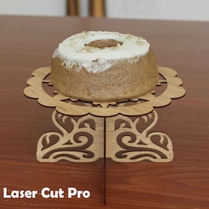 Puede incluir: Un soporte para pastel de madera con un diseño decorativo, que presenta una parte superior redonda y tres patas. Un pequeño pastel redondo con glaseado blanco está en la parte superior del soporte.