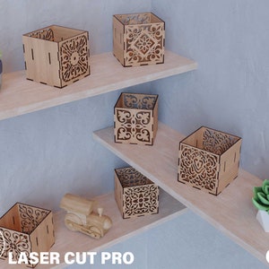 Puede incluir: Seis cajas decorativas de madera con intrincados diseños cortados con láser. Las cajas están dispuestas en dos estantes contra una pared gris claro. Las cajas son de diferentes tamaños y tienen un acabado de madera natural.