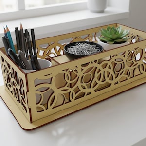 May include: A decorative wooden desk organizer with a laser-cut design. The organizer has compartments for pens, pencils, paperclips, and a small succulent. The organizer is light brown with a darker brown trim. The text "Laser Cut Pro" is in the lower left corner.