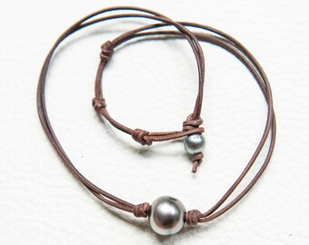 May include: A brown leather cord necklace with two dark gray pearls. The necklace is adjustable and can be worn at different lengths.