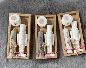 May include: Three wooden boxes each contain a white candle holder, a white nutcracker figurine, and a candle with "Frohes Fest" text. The candles are in shades of green, grey, and white. Each box is decorated with dried flowers and red and white twine.