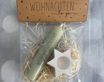 May include: A festive gift set in a clear bag, featuring a pale green candle inscribed with "WEIHNACHTSLICHT" and a white star-shaped candle holder. The set is presented with straw and a brown cardboard tag with the text "WEIHNACHTEN to go".