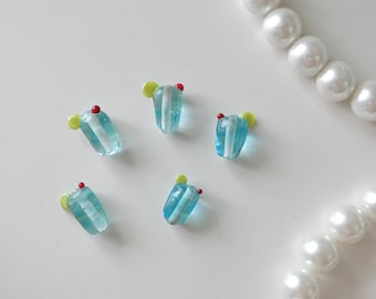 May include: Five small, translucent blue glass beads shaped like drinking glasses, each featuring a white vertical stripe, a tiny yellow accent, and a small red dot. A string of white, round, lustrous beads is positioned in the upper right corner.