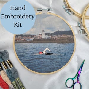 May include: Hand embroidery kit featuring a swimmer in the sea, with a lighthouse and town in the background. The kit includes embroidery floss, a wooden hoop, and scissors. The text "Hand Embroidery Kit" is in a blue circle.