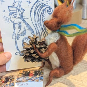 May include: A handcrafted felt squirrel figurine with a pinecone, a blue and green scarf, and a blue ink drawing of a squirrel. The squirrel is brown with a white chest. Business cards are visible in the foreground.