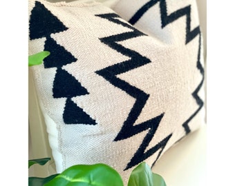 May include: Cream cushion with a black geometric zigzag pattern. The cushion has a textured weave and a soft appearance. The design features bold, contrasting black shapes against a neutral background, adding a modern touch to home decor.