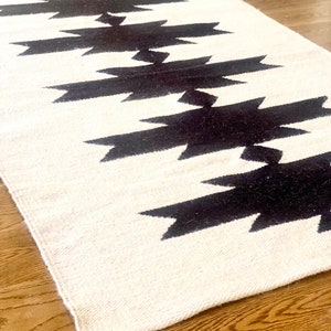 May include: A white and black geometric rug with a repeating pattern of black arrow shapes. The rug is made of a soft, woven material and has a fringed edge.