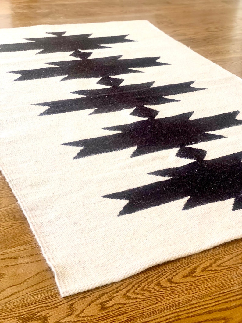 May include: A white and black geometric rug with a repeating pattern of black arrow shapes. The rug is made of a soft, woven material and has a fringed edge.