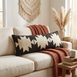 Könnte beinhalten: Ein rechteckiges Lendenkissen mit einem geometrischen Muster in Schwarz und Creme. Das Kissen liegt auf einem hellen Sofa neben einer strukturierten, rostfarbenen Wohndecke, was einen Kontrast zwischen dem dunklen Stoff und dem neutralen Bezug erzeugt.