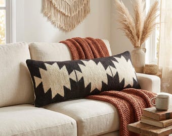 Puede incluir: Un cojín lumbar rectangular con un patrón geométrico en negro y crema. El cojín está colocado sobre un sofá de color claro junto a una manta texturizada de color óxido, creando un contraste entre la tela oscura y la tapicería neutra.