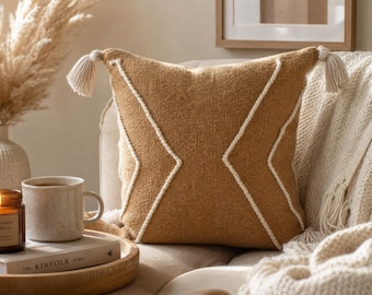 May include: A square throw pillow in a warm tan color featuring a textured woven fabric, white corded geometric chevron patterns, and small white tassels attached to the two top corners. The pillow is displayed on a soft, neutral-toned sofa.