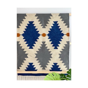 May include: A rectangular wall hanging rug with a geometric pattern. The rug features a repeating design of blue, grey, and cream-coloured diamond shapes. The rug is approximately 122 cm wide and 152 cm long, with a fringe along the bottom edge.