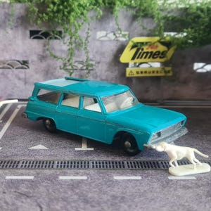 Könnte beinhalten: Ein türkisfarbener Spielzeugkombi mit weißem Innenraum, ein weißer Hund und eine weiße Figur. Eine Matchbox-Box befindet sich links. Die Szene spielt sich auf einer grauen Oberfläche mit weißen Linien ab, mit einem Times-Schild im Hintergrund.