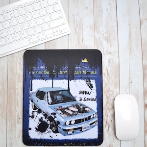 May include: A rectangular mousepad with a light blue BMW 3 Series car image, hood open. The mousepad has a blue background with black paint splatter and text reading "Haynes Se... and repair Manual" and "BMW 3 Series". A white keyboard and mouse are visible.