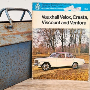 May include: A blue metal toolbox with a rusty patina sits next to a vintage car repair manual for Vauxhall Velox, Cresta, Viscount, and Ventora models. The manual is from the RAC (Royal Automobile Club) series for owner drivers and is approved and recommended. The manual cover features a white car in a park setting with trees and fallen leaves.