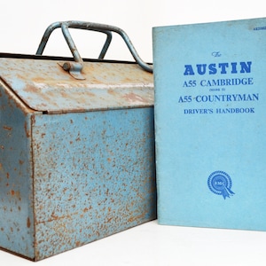 Op de afbeelding: Een blauwe metalen gereedschapskist met een roestig oppervlak en een handvat. Ernaast ligt een blauw boekje met de titel "The Austin A55 Cambridge Mark II A55 Countryman Driver's Handbook" met een BMC-logo.