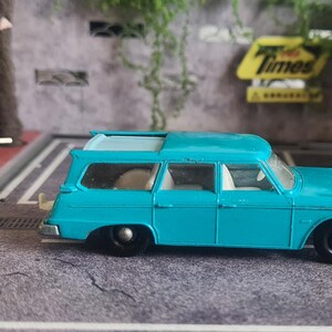 May include: A turquoise toy estate car with black wheels, a white dog figurine, and a white figurine of a person with a gun. A Matchbox box is in the background.