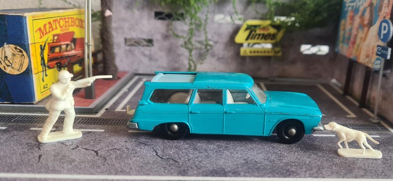 May include: A turquoise toy estate car with black wheels, a white dog figurine, and a white figurine of a person with a gun. A Matchbox box is in the background.