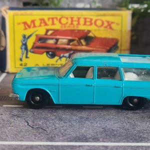 May include: A vintage Matchbox toy set featuring a turquoise station wagon, a white figure aiming a rifle, and a white dog figurine. A yellow Matchbox box is in the background.