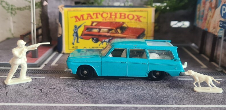 May include: A vintage Matchbox toy set featuring a turquoise station wagon, a white figure aiming a rifle, and a white dog figurine. A yellow Matchbox box is in the background.
