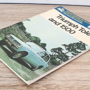 May include: A blue Triumph Toledo 1500 car driving on a road with trees on either side. The car has a white license plate that reads "HMC 450H". The image is from a vintage RAC (Royal Automobile Club) service manual.