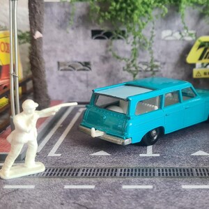 May include: A miniature scene with a light blue toy station wagon, a white toy dog, and a white toy figure. A vintage Matchbox box is visible. The scene is set on a grey surface with painted lines, resembling a car park.