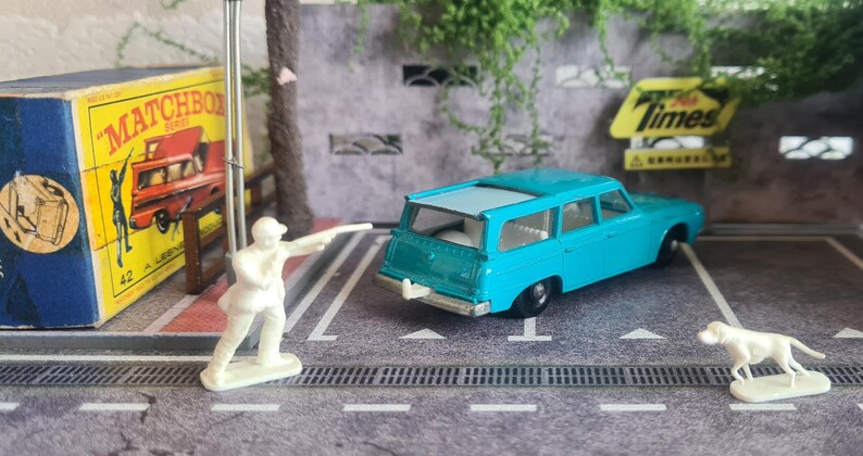May include: A miniature scene with a light blue toy station wagon, a white toy dog, and a white toy figure. A vintage Matchbox box is visible. The scene is set on a grey surface with painted lines, resembling a car park.