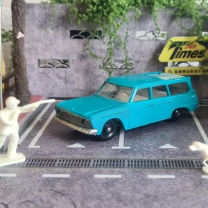 May include: A turquoise station wagon toy with black wheels, parked on a grey surface. A white figurine of a person and a dog are nearby. A vintage Matchbox box and a yellow sign with the word "Times" are in the background.