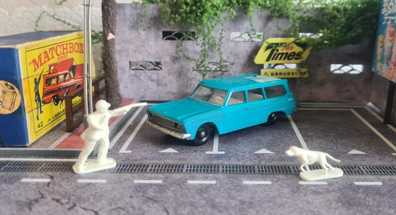 May include: A turquoise station wagon toy with black wheels, parked on a grey surface. A white figurine of a person and a dog are nearby. A vintage Matchbox box and a yellow sign with the word "Times" are in the background.