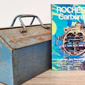Puede incluir: Una caja de herramientas de metal azul con óxido y un libro titulado "Carburadores Rochester" con un diagrama de un carburador en la portada.