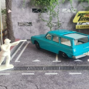 May include: A teal miniature estate car, a white figurine of a person pointing, and a white dog figurine are on a grey surface. A vintage Matchbox box and a yellow sign with the word "Times" are in the background.