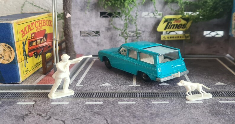 May include: A teal miniature estate car, a white figurine of a person pointing, and a white dog figurine are on a grey surface. A vintage Matchbox box and a yellow sign with the word "Times" are in the background.
